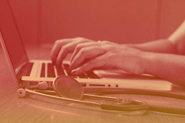 A stethoscope lying a table next to a person using a laptop